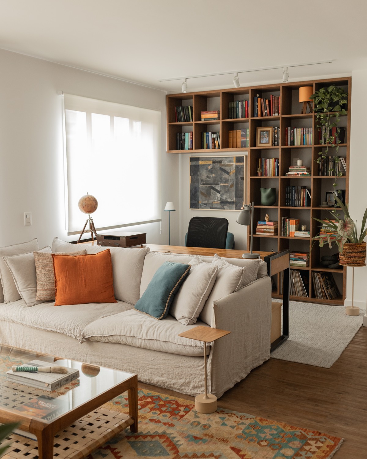Sala integrada com estante de madeira, sofá claro e home office ao fundo