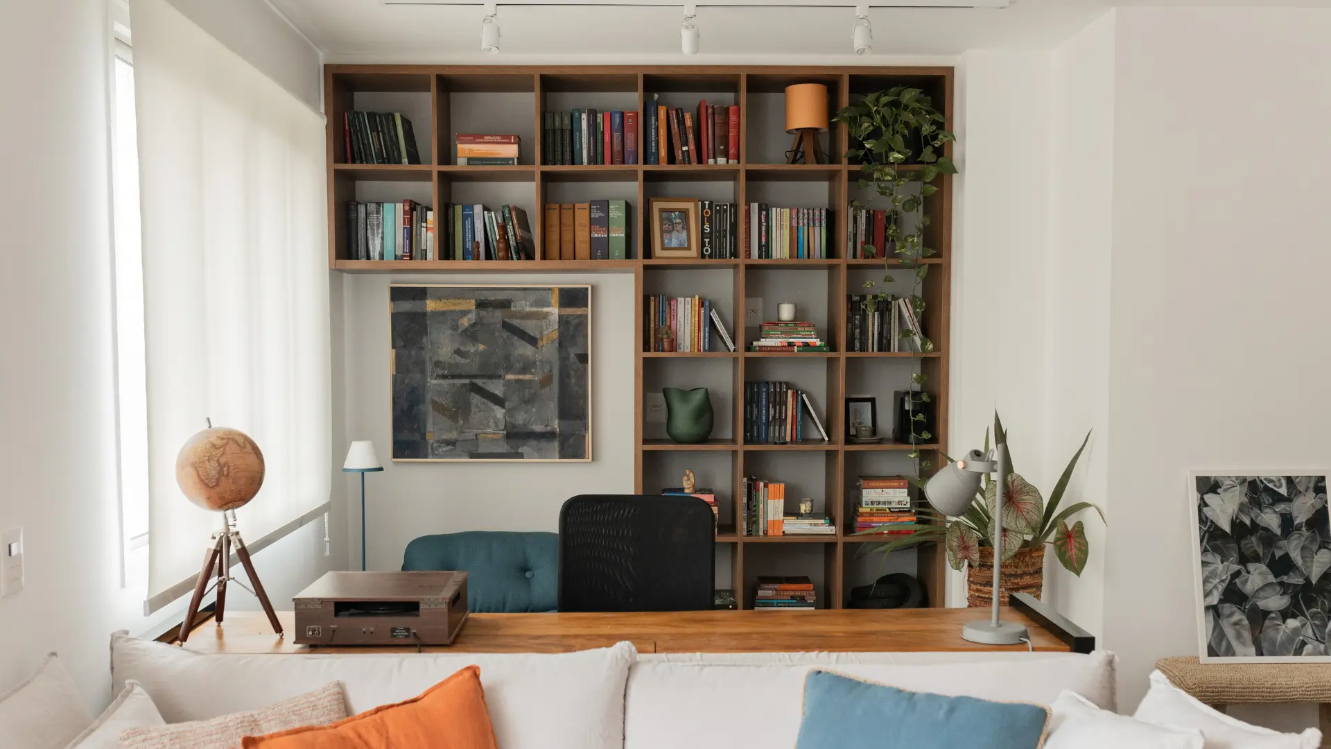 Home office pronto, com estante de madeira cheia de livros, bancada atrás do sofá e almofadas coloridas