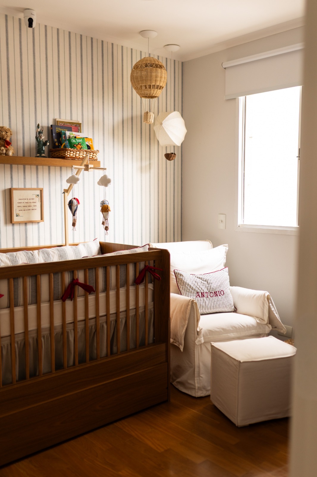 Quarto de bebê com berço de madeira, poltrona clara e papel de parede listrado